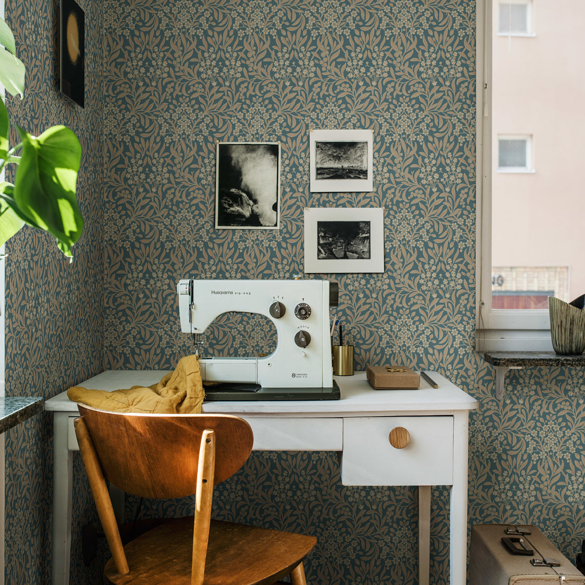 Vintage Husqvarna sewing machine on a white desk with mustard cloth, wooden chair, teal floral wallpaper and framed photos.