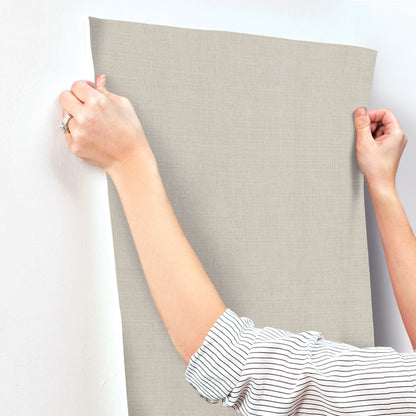 Hands stretch a gray linen canvas onto a plain wall; striped shirt sleeves visible.