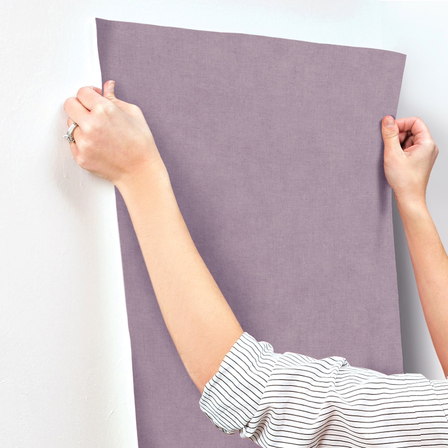 Two hands hang a lavender faux linen wallpaper panel against a wall.