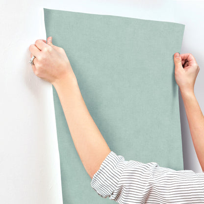 Rectangular sage-green fabric panel being stretched by hands; striped-sleeve person against a white wall.