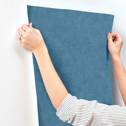 Blue denim-like faux linen wallpaper panel held by two hands against a white wall; striped-sleeve arm visible.