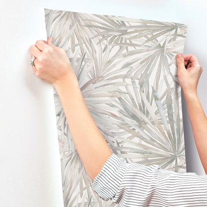 Two hands hold a rectangular wallpaper sample with a gray palm-leaf pattern; white background, striped sleeves visible.