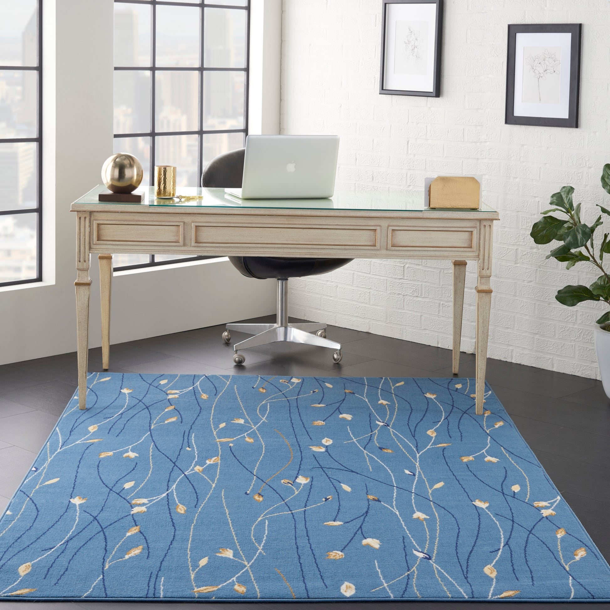 Cream carved-wood desk with glass top in a bright office, chrome swivel chair, blue abstract rug, laptop and gold decor.