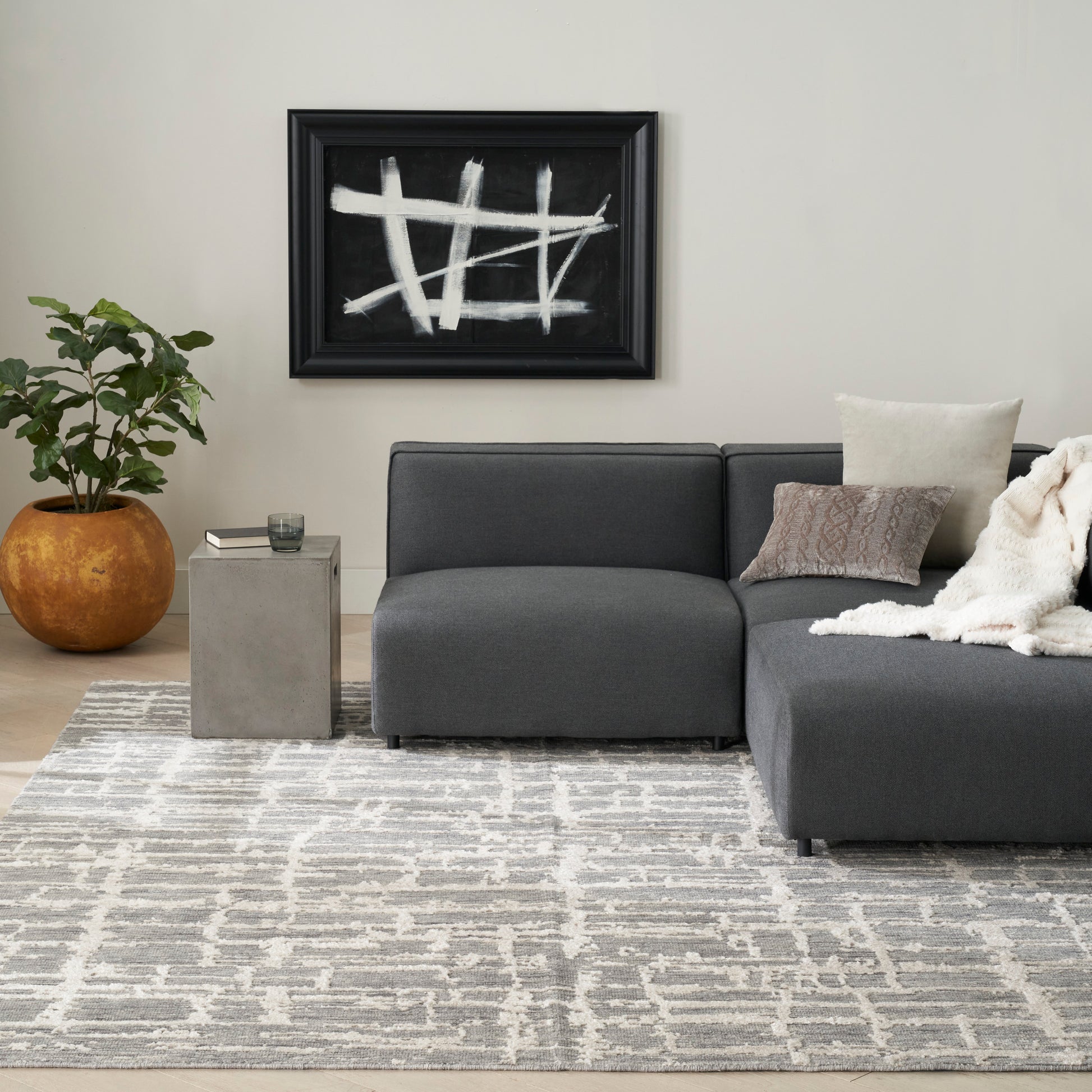 Dark gray fabric sectional with chaise, beige pillows and throw, concrete side table, geometric gray rug, black abstract art.