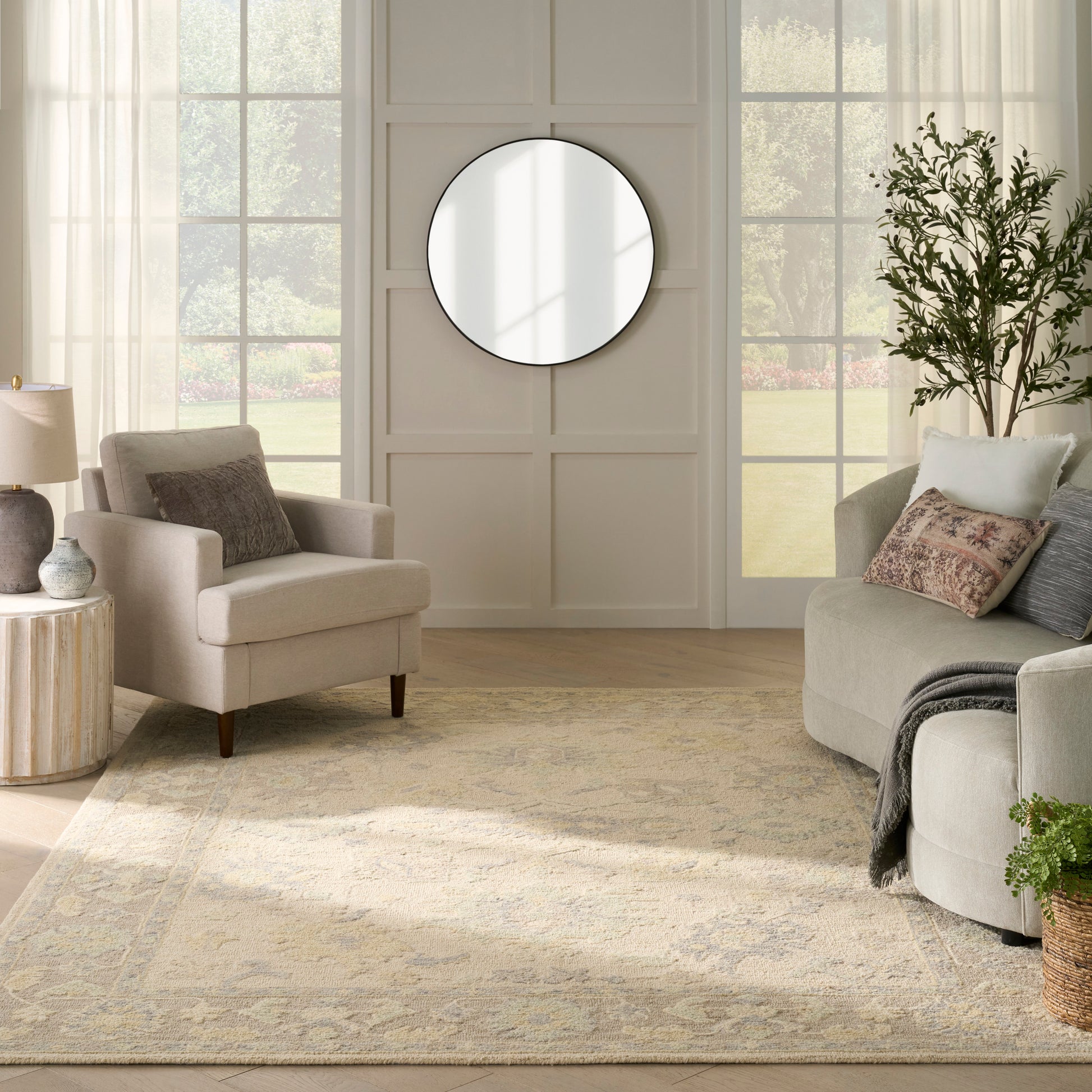 Neutral living room with beige/gray seating, round wall mirror, light wood floor, patterned rug, plant, and sheer curtains.