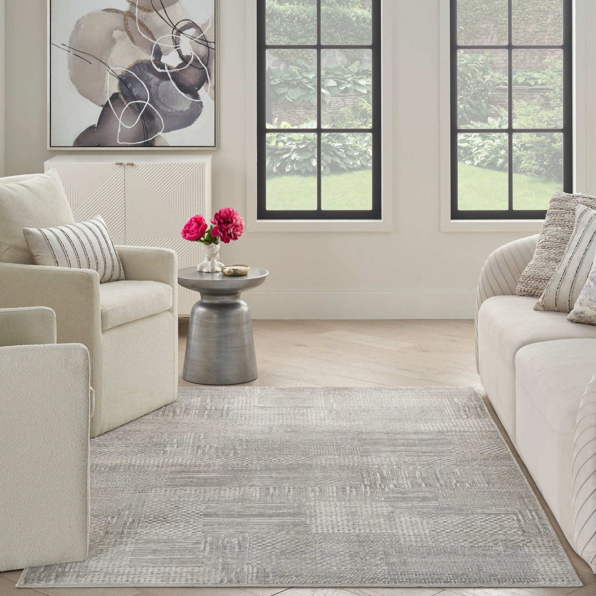 Light gray geometric area rug in a beige living room with cream sofas, a metal side table, and large black-framed windows.