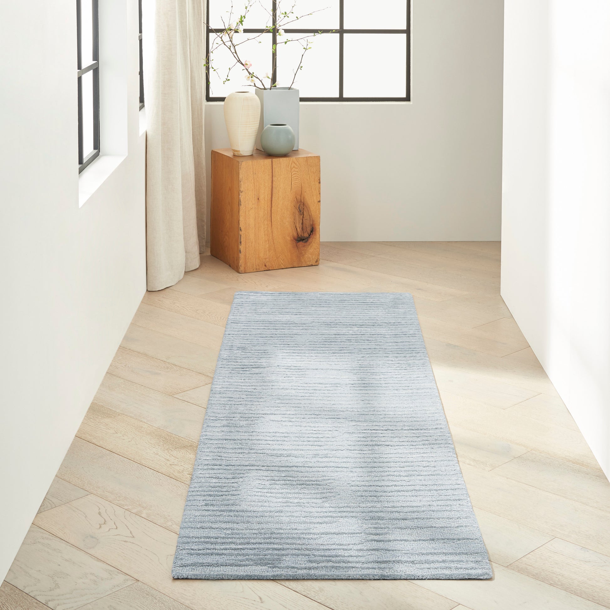 Light blue-gray striped runner rug along a pale wood floor in a bright hallway with a wooden block and vases.