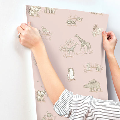 Hands applying pale pink wallpaper featuring beige wildlife drawings (giraffes, elephants, penguin).