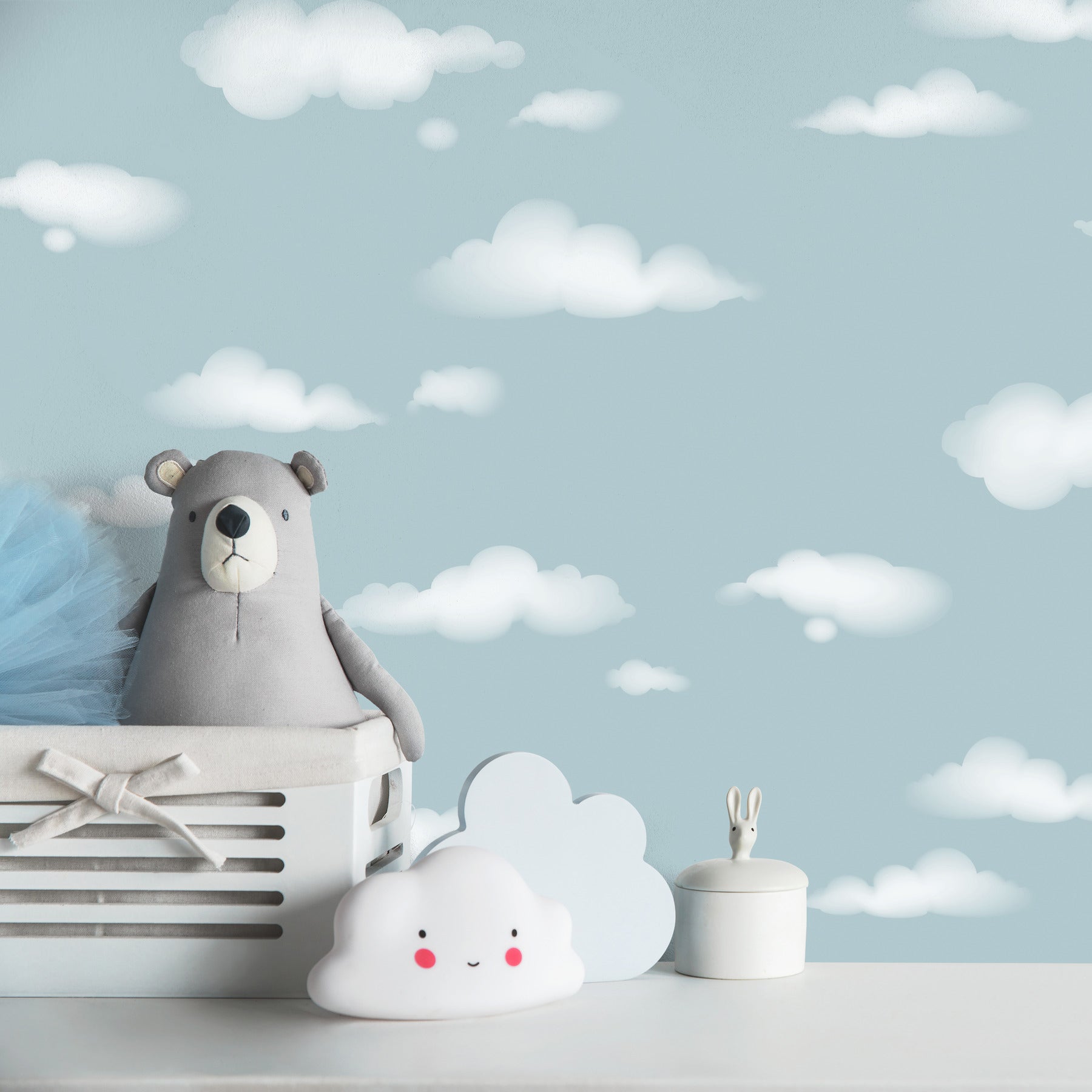 Grey plush bear in a white crate with ribbon, cloud plush and bunny-ear jar against a blue nursery cloud backdrop.