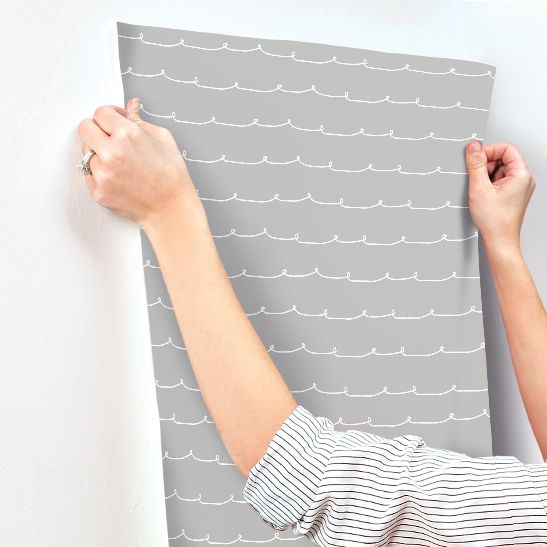 Grey wallpaper panel with white wavy lines being applied to a wall, hands in striped sleeves holding it