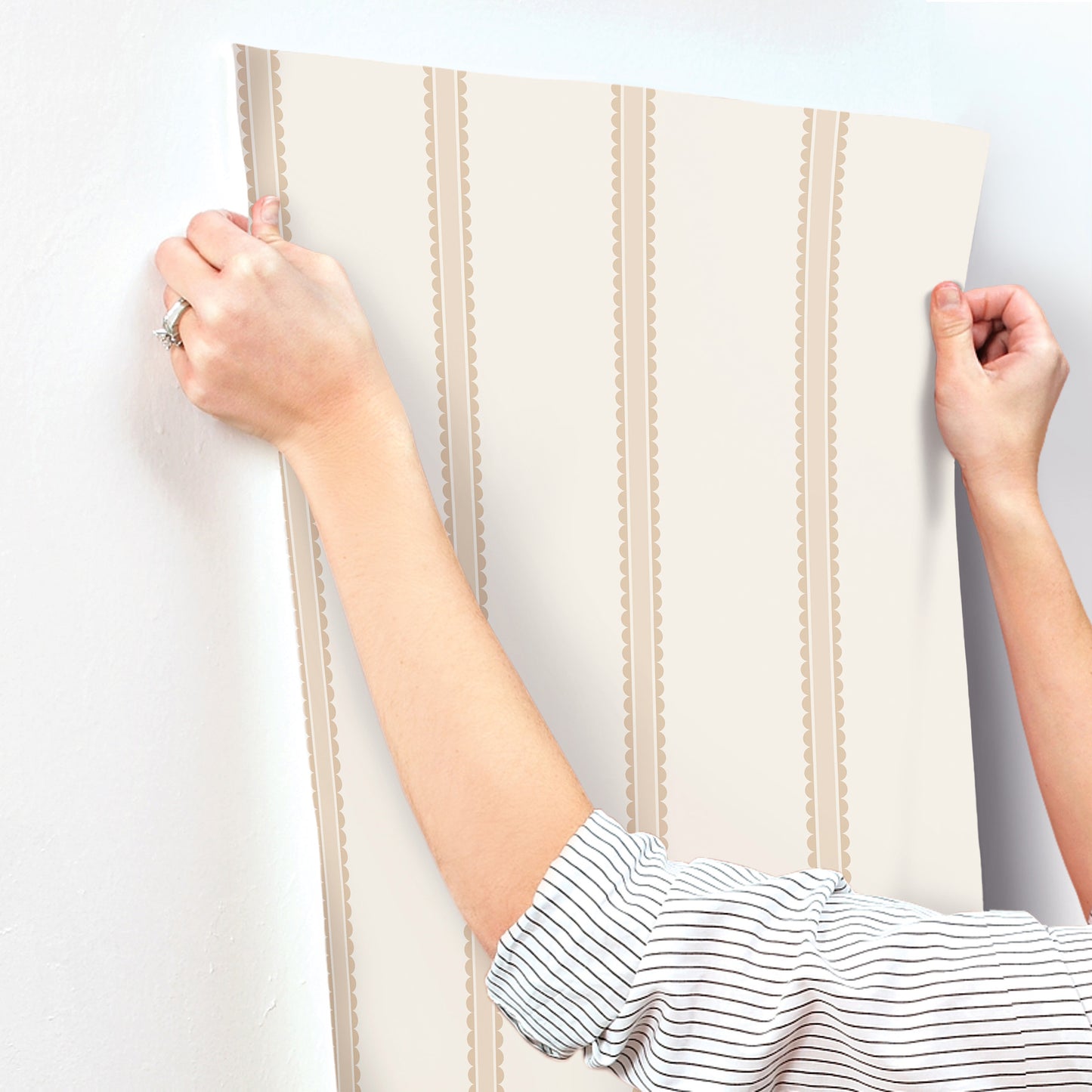 Beige wallpaper panel with vertical brown scalloped stripes, held by two hands as if to hang.