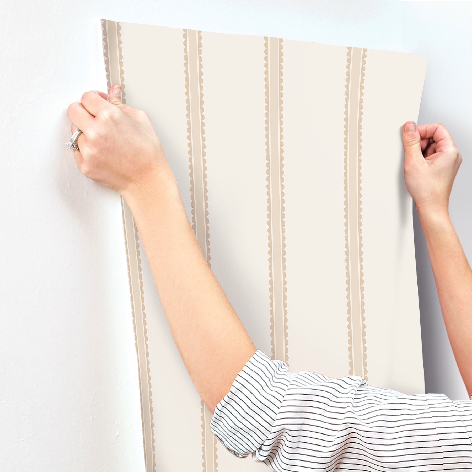 Beige wallpaper panel with vertical brown scalloped stripes, held by two hands as if to hang.