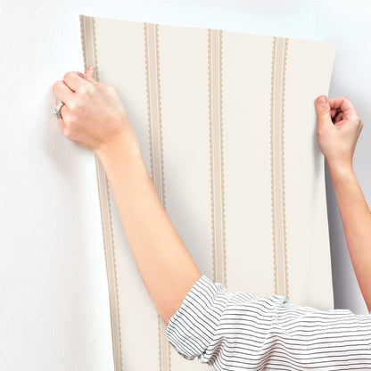 Beige wallpaper panel with vertical brown scalloped stripes, held by two hands as if to hang.