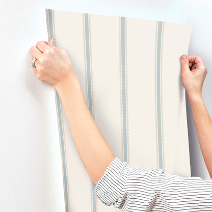 Beige wallpaper panel with pale blue vertical stripes and dotted trim, held by two hands.