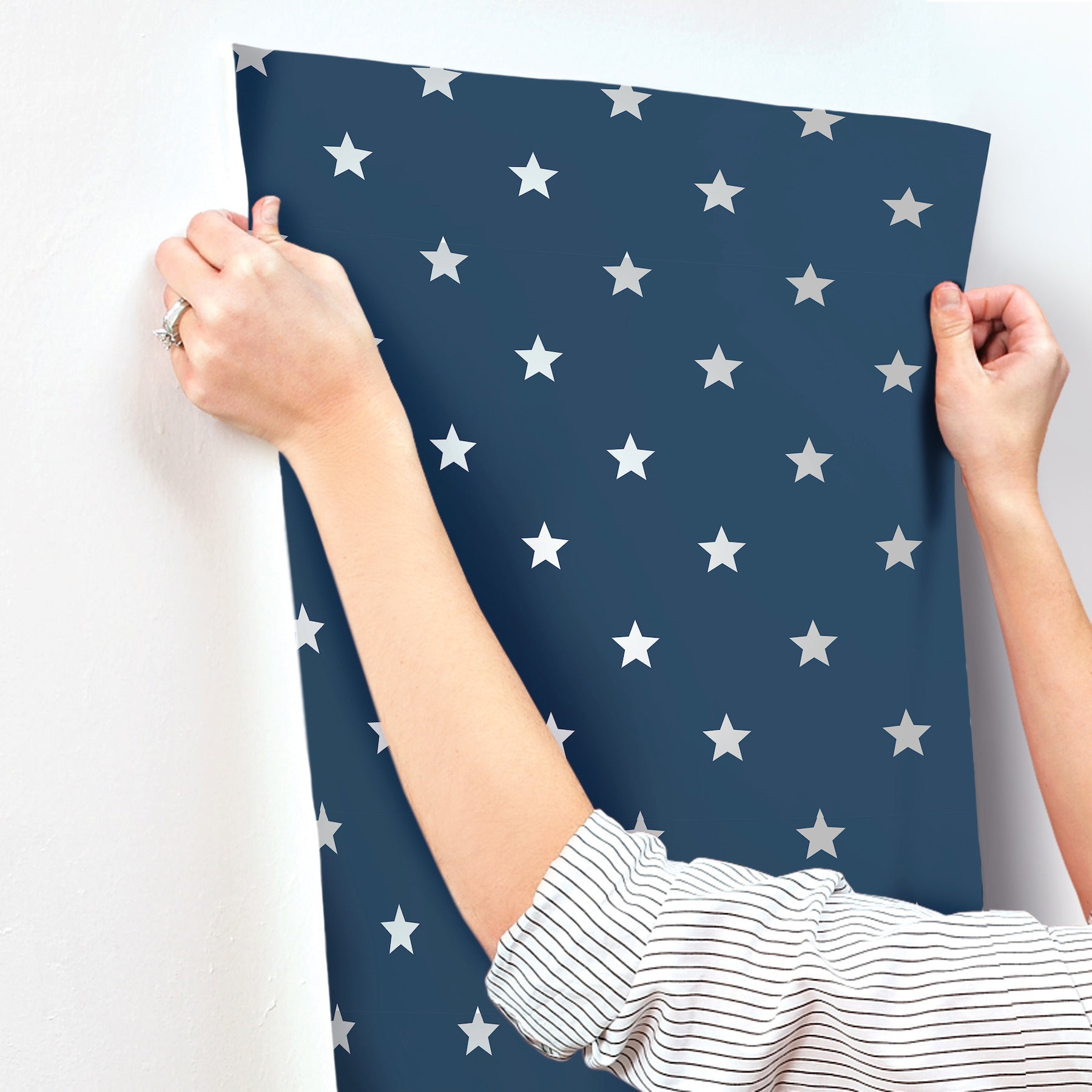 Hands hold a navy blue wallpaper sheet with white stars; striped sleeve visible, curved edge against wall.