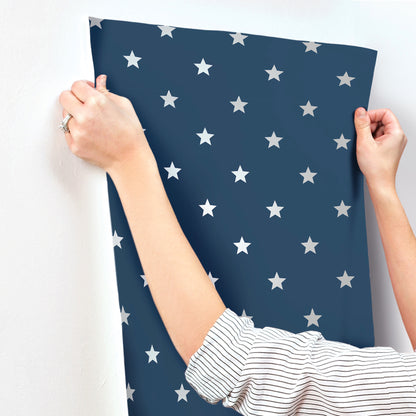 Hands hold a navy blue wallpaper sheet with white stars; striped sleeve visible, curved edge against wall.