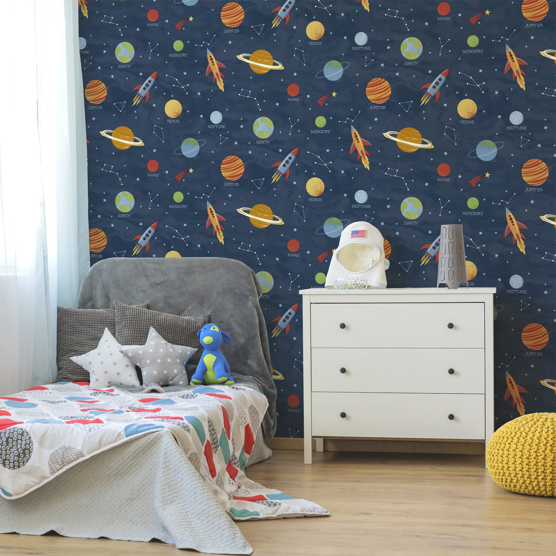Kids' room with blue outer space wallpaper, white dresser, grey upholstered bed, star pillows, plush astronaut, and yellow pouf.