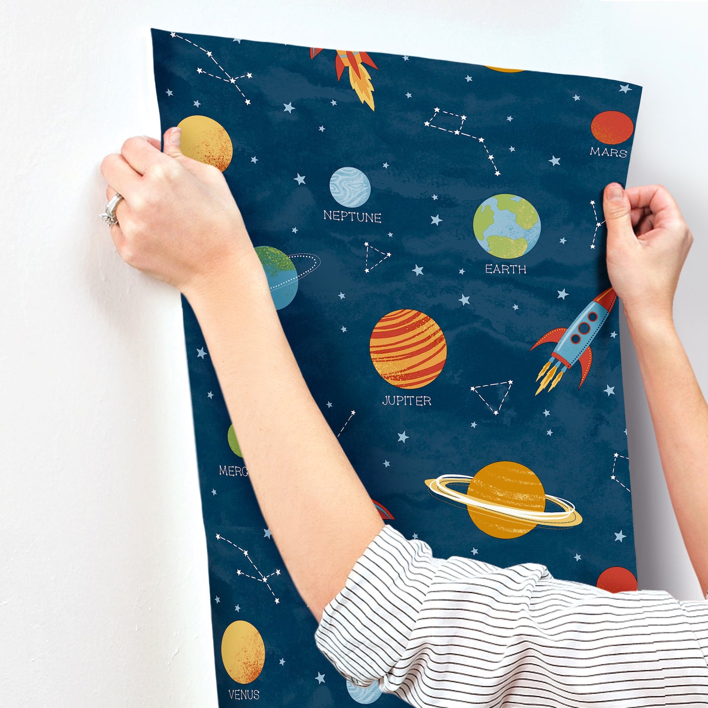Dark blue space-themed fabric featuring labeled planets, a rocket, and constellations; hands pull at the edges.