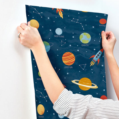 Dark blue space-themed fabric featuring labeled planets, a rocket, and constellations; hands pull at the edges.