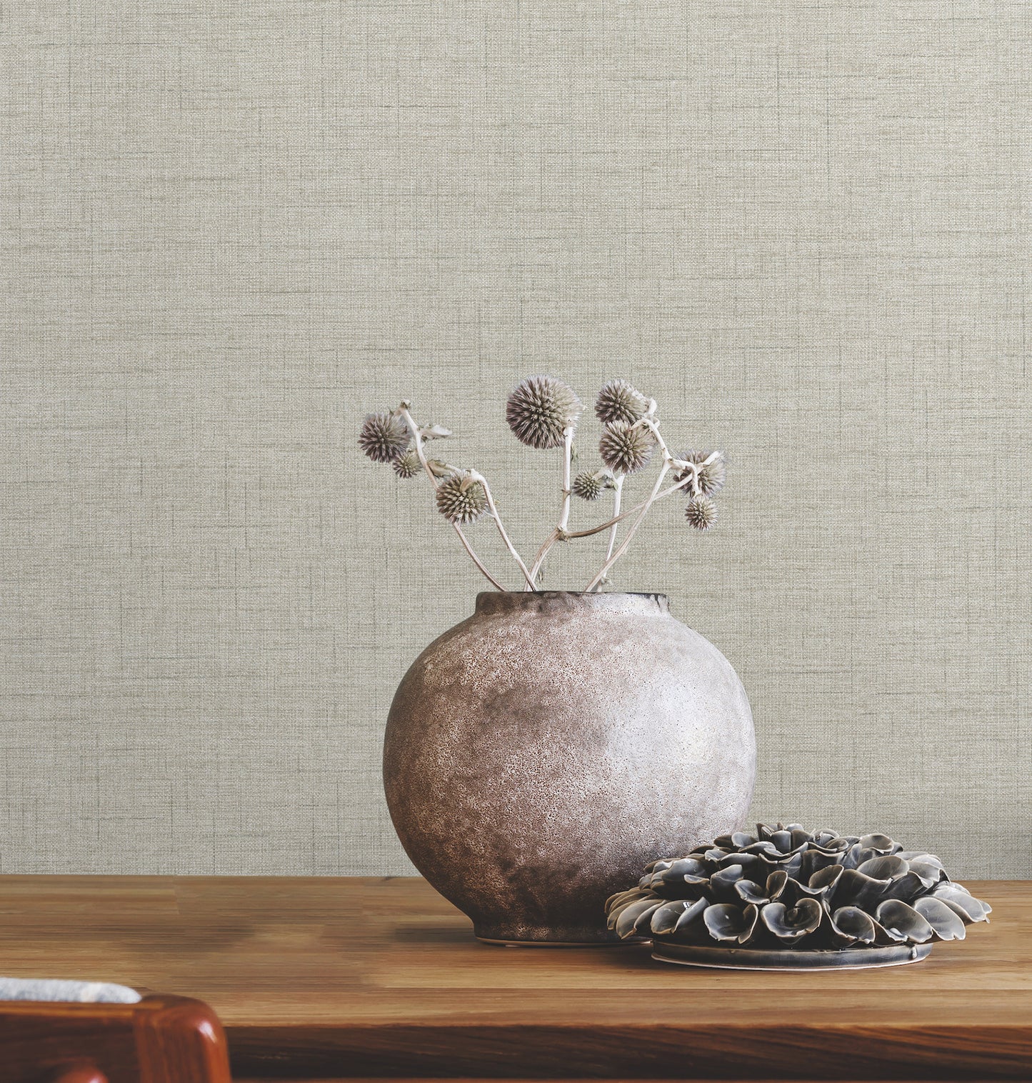 Round ceramic vase with dried thistles on a wooden table, beside a plate of dark shells, beige textured wall backdrop.