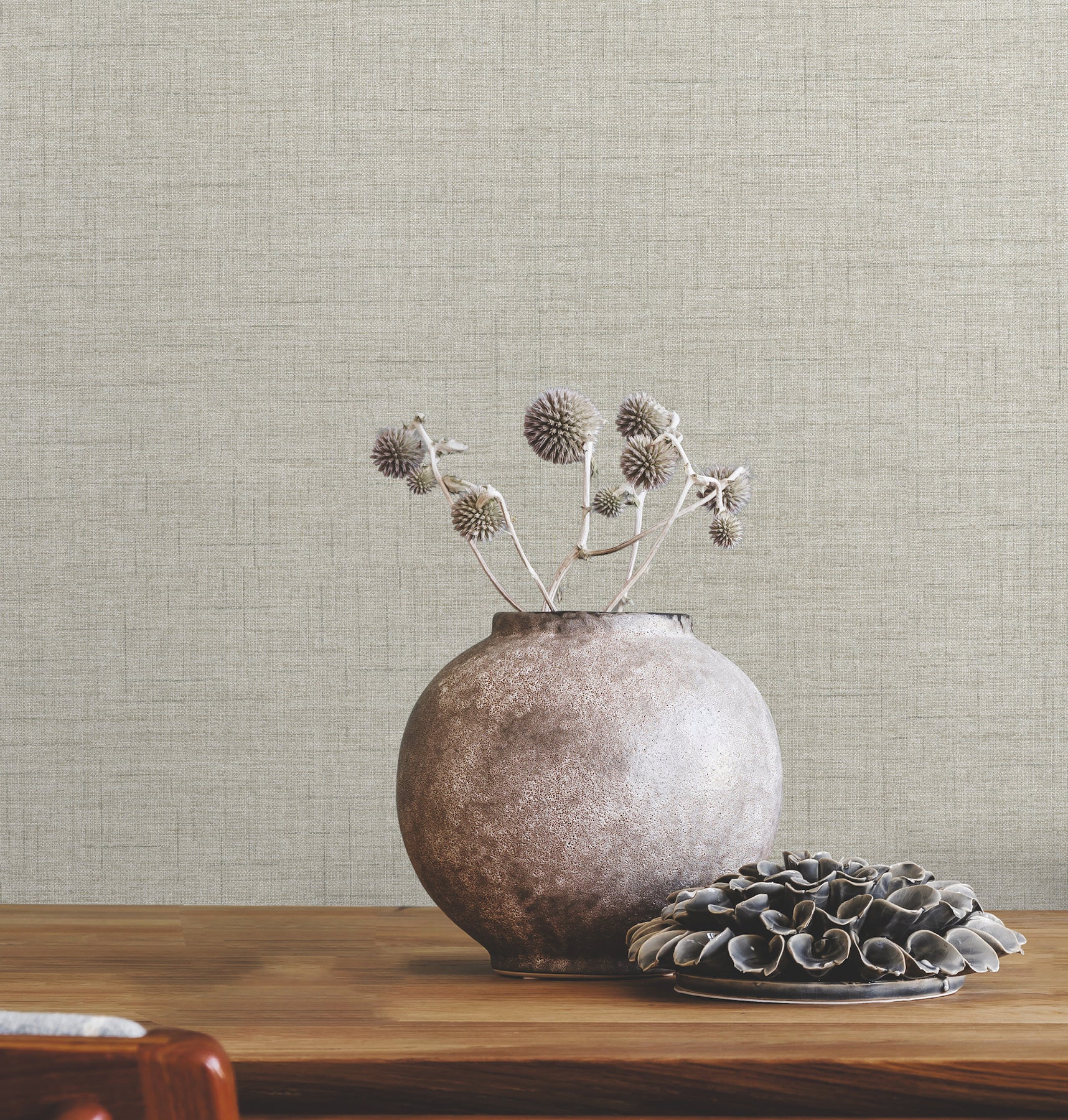 Round ceramic vase with dried thistles on a wooden table, beside a plate of dark shells, beige textured wall backdrop.