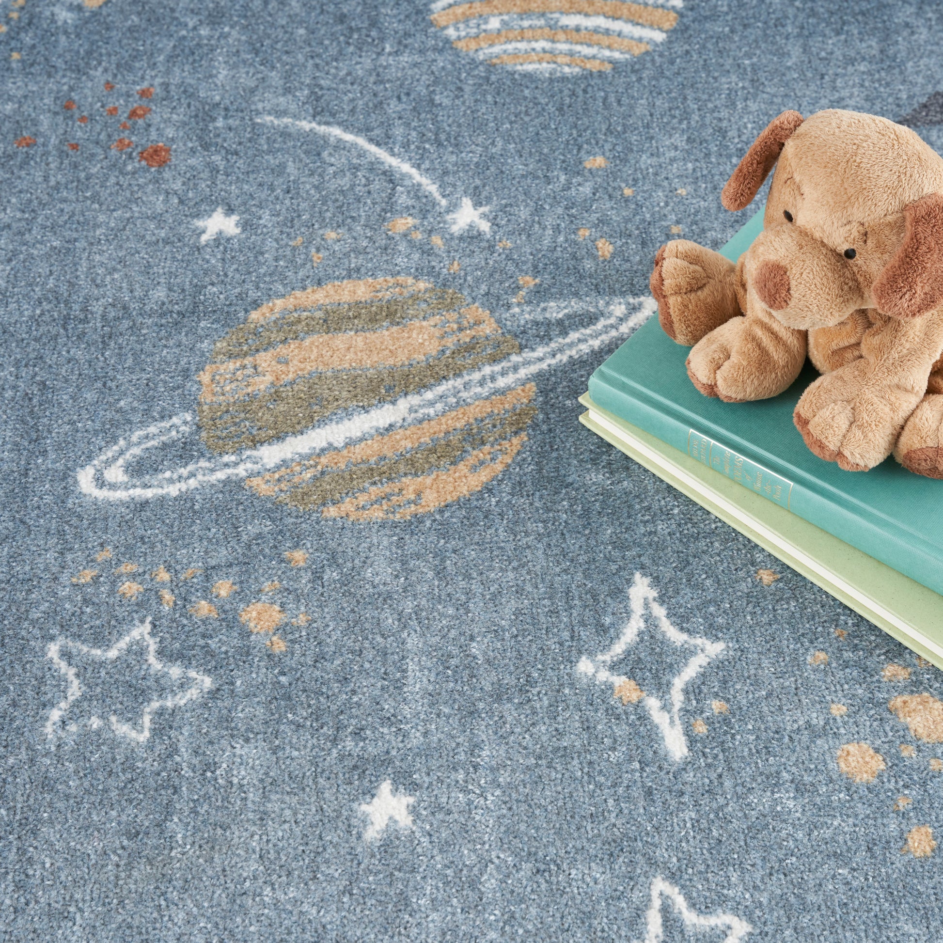 Blue space rug with Saturn-like planet and stars; tan plush dog sits on green and turquoise books.