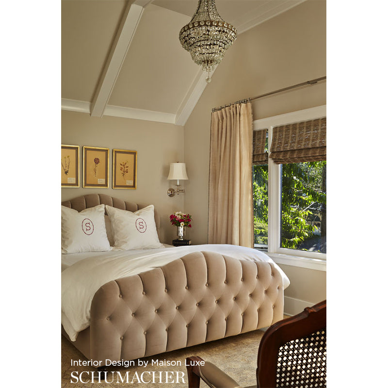 Beige tufted upholstered bed in a chandelier-lit bedroom with framed art, lamp, and a window with curtains.