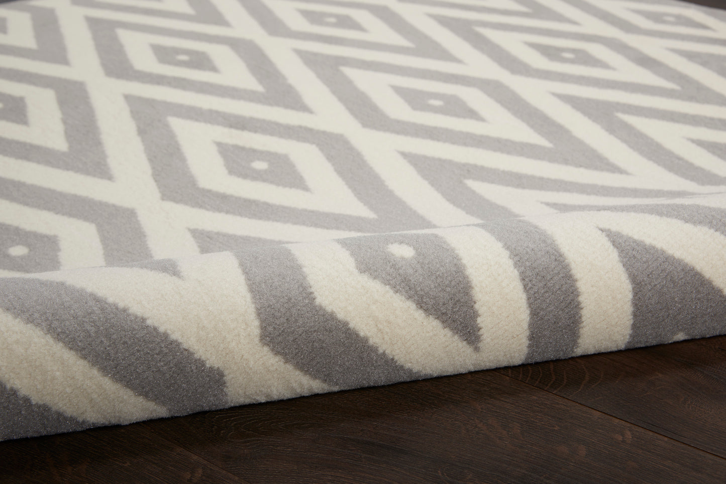 Grey round rug with white geometric diamond pattern, rolled edge, plush pile on dark wood floor.