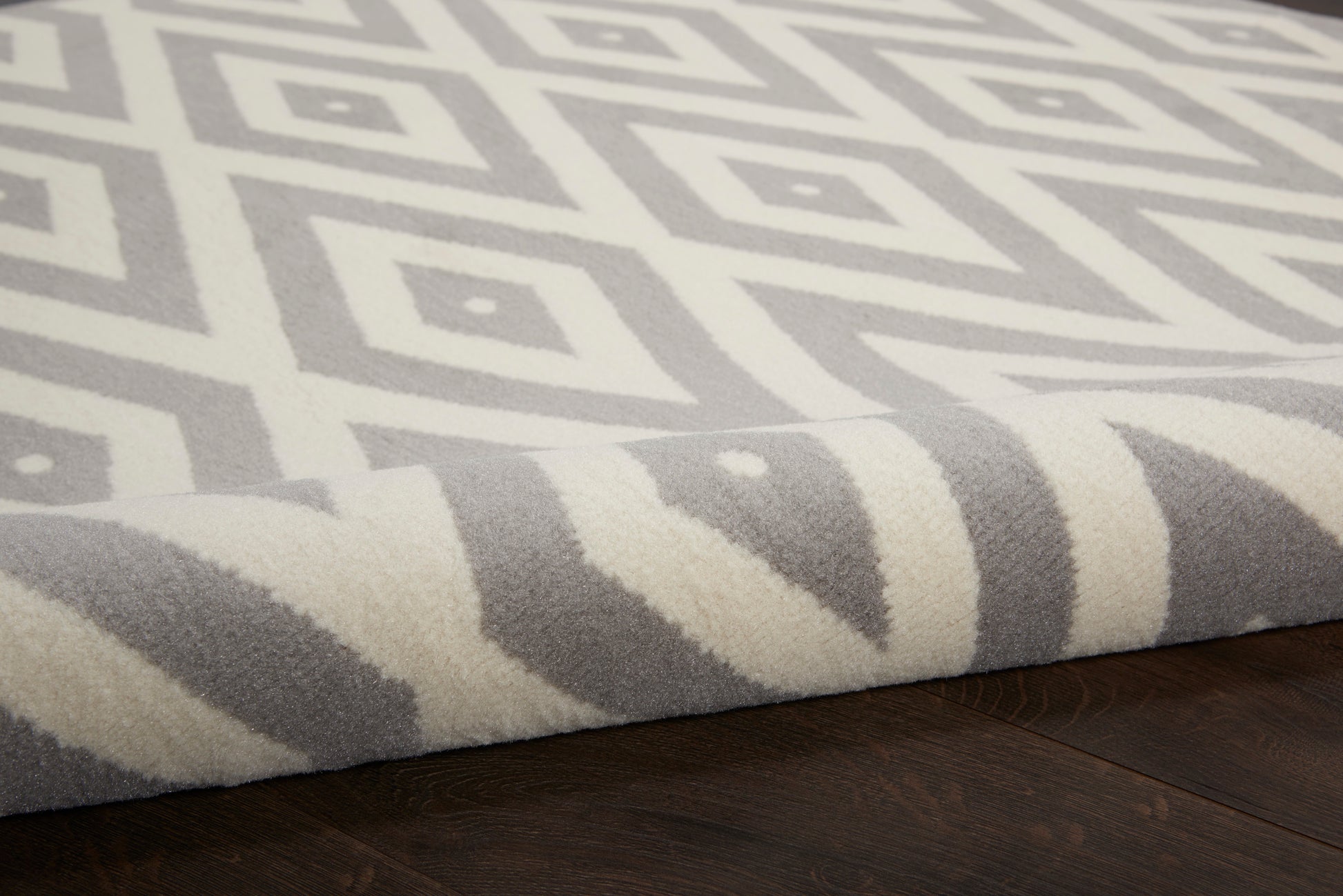 Grey round rug with white geometric diamond pattern, rolled edge, plush pile on dark wood floor.