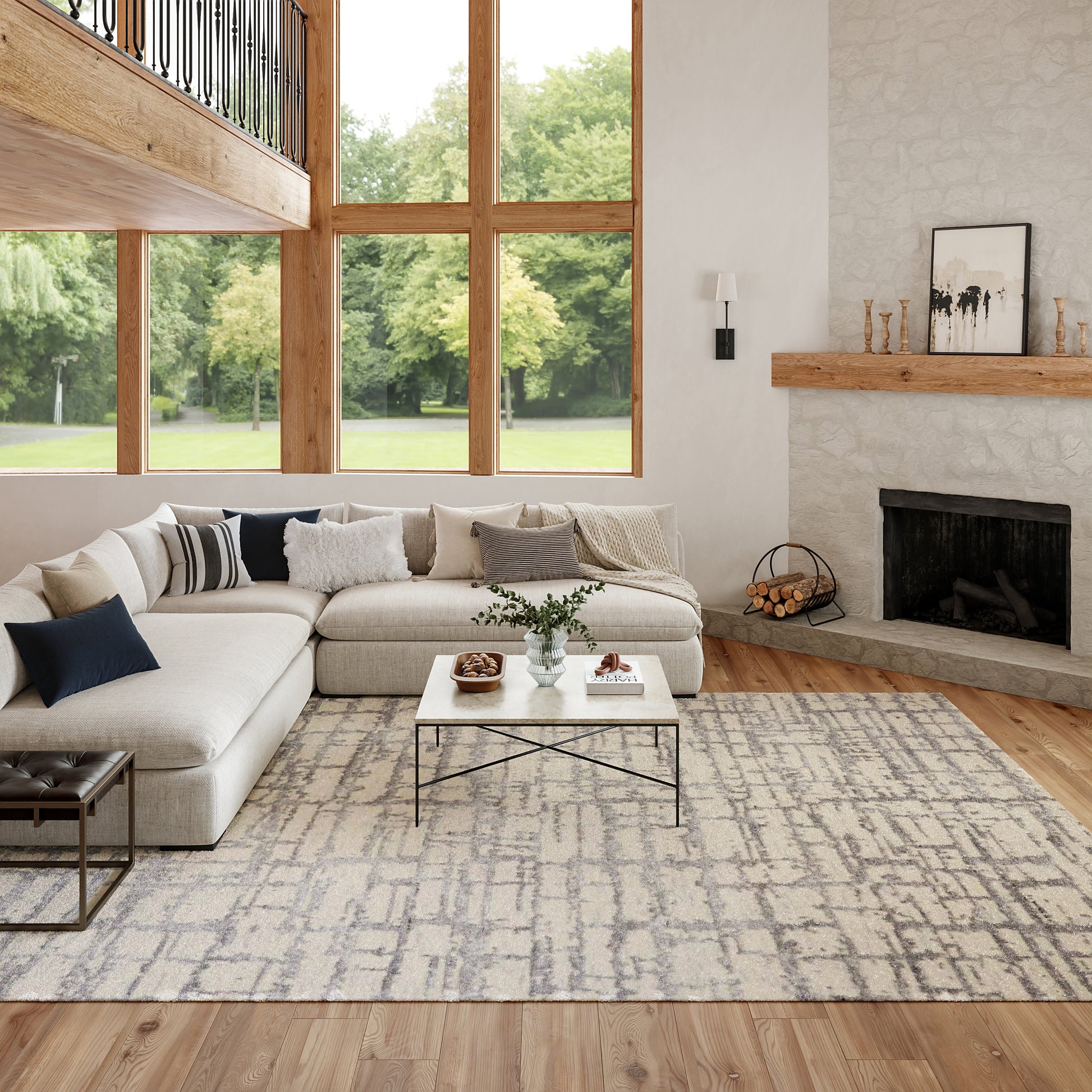 Bright living room with beige sectional, grey rug, large wood-framed windows, and a stone fireplace with a wooden mantle.