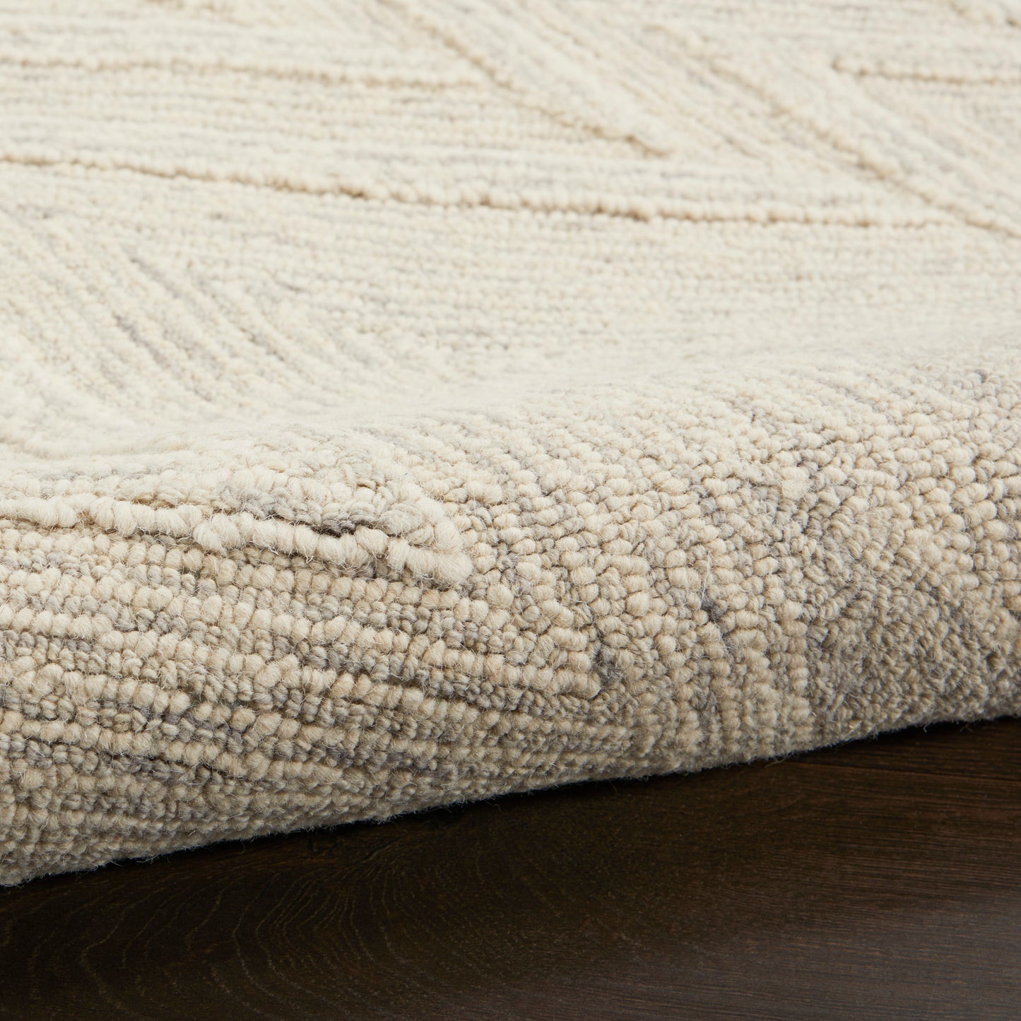 Cream textured rug with loop pile and subtle geometric pattern, edge resting on dark wood floor.