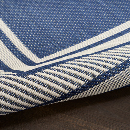 Blue woven rug with white striped border and twisted fringe edge on dark wood floor.
