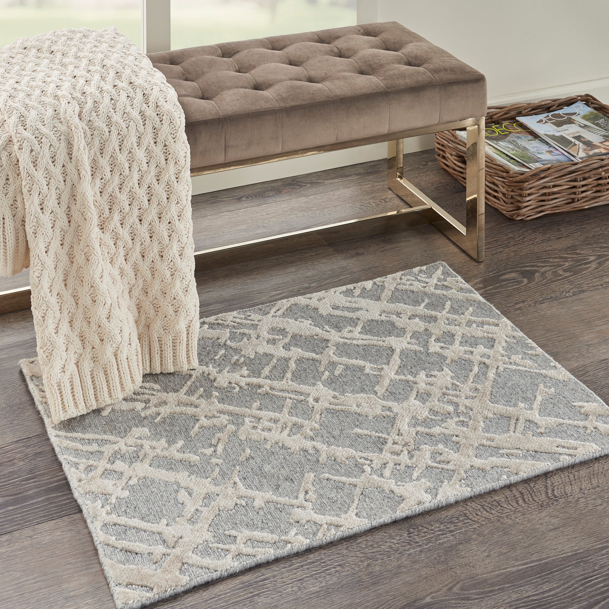 Taupe tufted bench with gold metal frame, cream knit throw, grey lattice rug, and wicker magazine rack on dark wood floor.