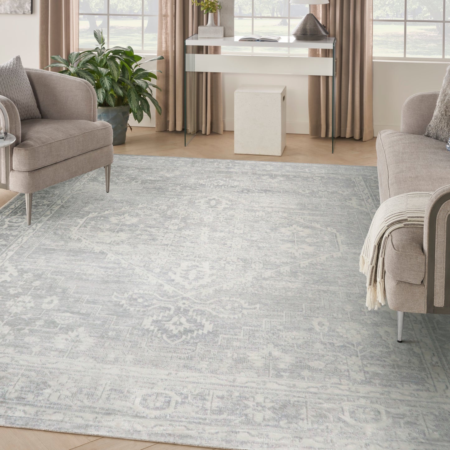 Pale blue-gray patterned rug anchors a beige living room with upholstered chairs, light wood floors, and a white console near windows.