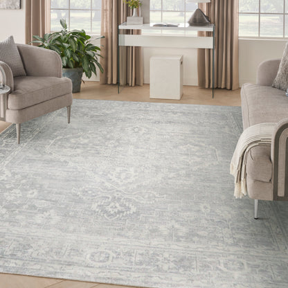 Pale blue-gray patterned rug anchors a beige living room with upholstered chairs, light wood floors, and a white console near windows.