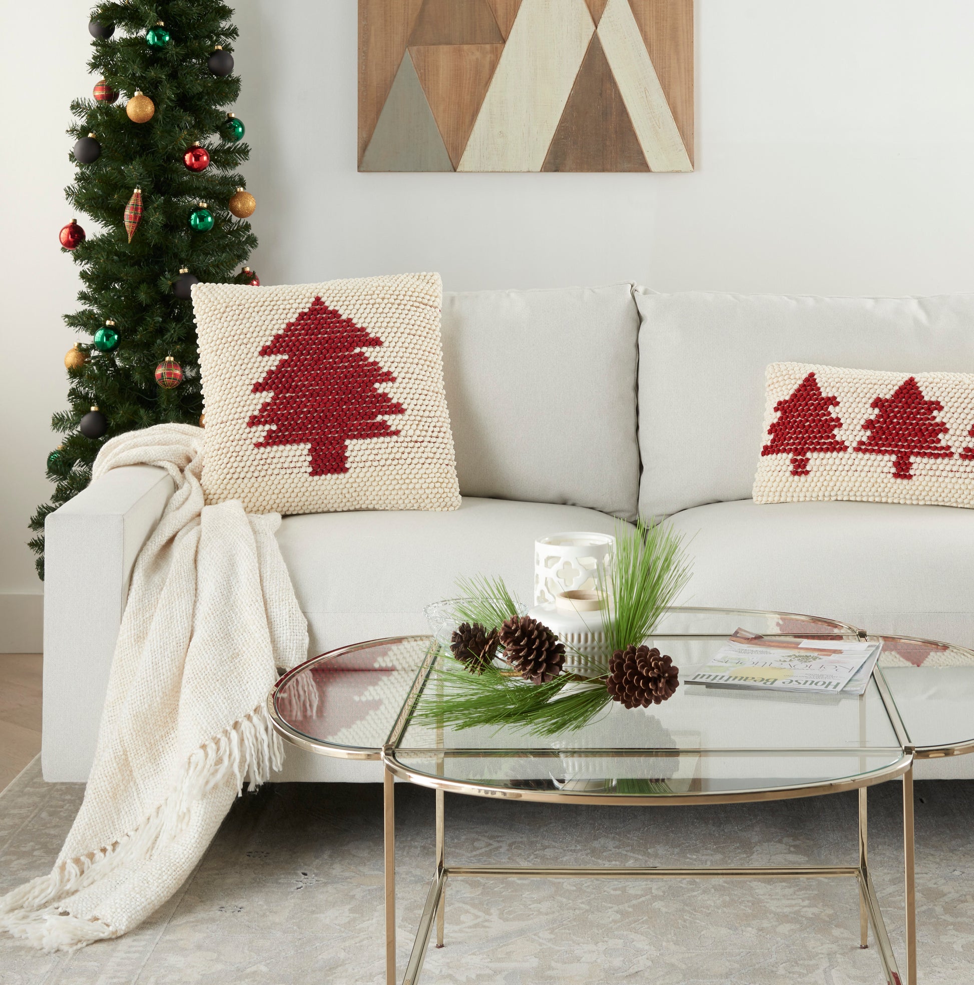 Cream sofa with red tree knitted pillows, beige throw, glass coffee table with pinecones, Christmas tree and wall art.