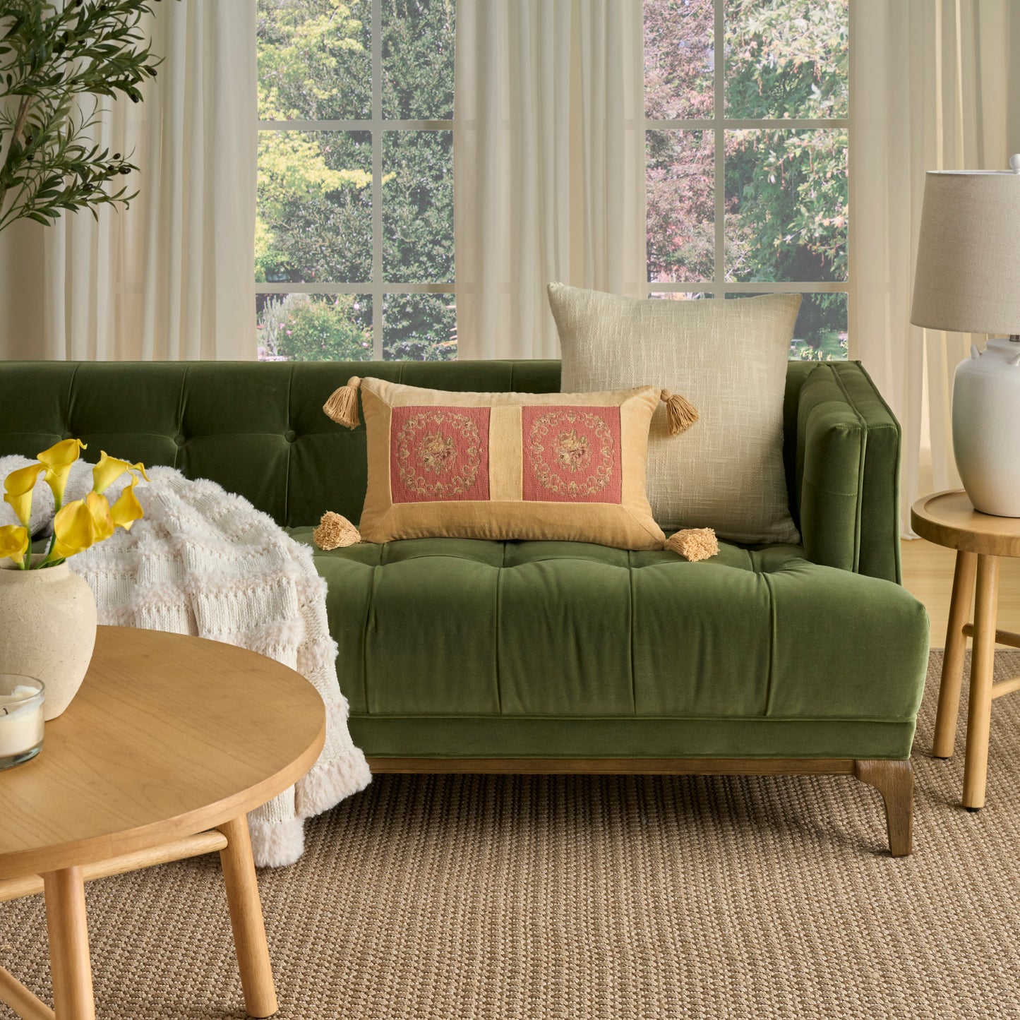 Olive green velvet tufted sofa with beige tassel pillows in a living room; round wood coffee table with yellow flowers.