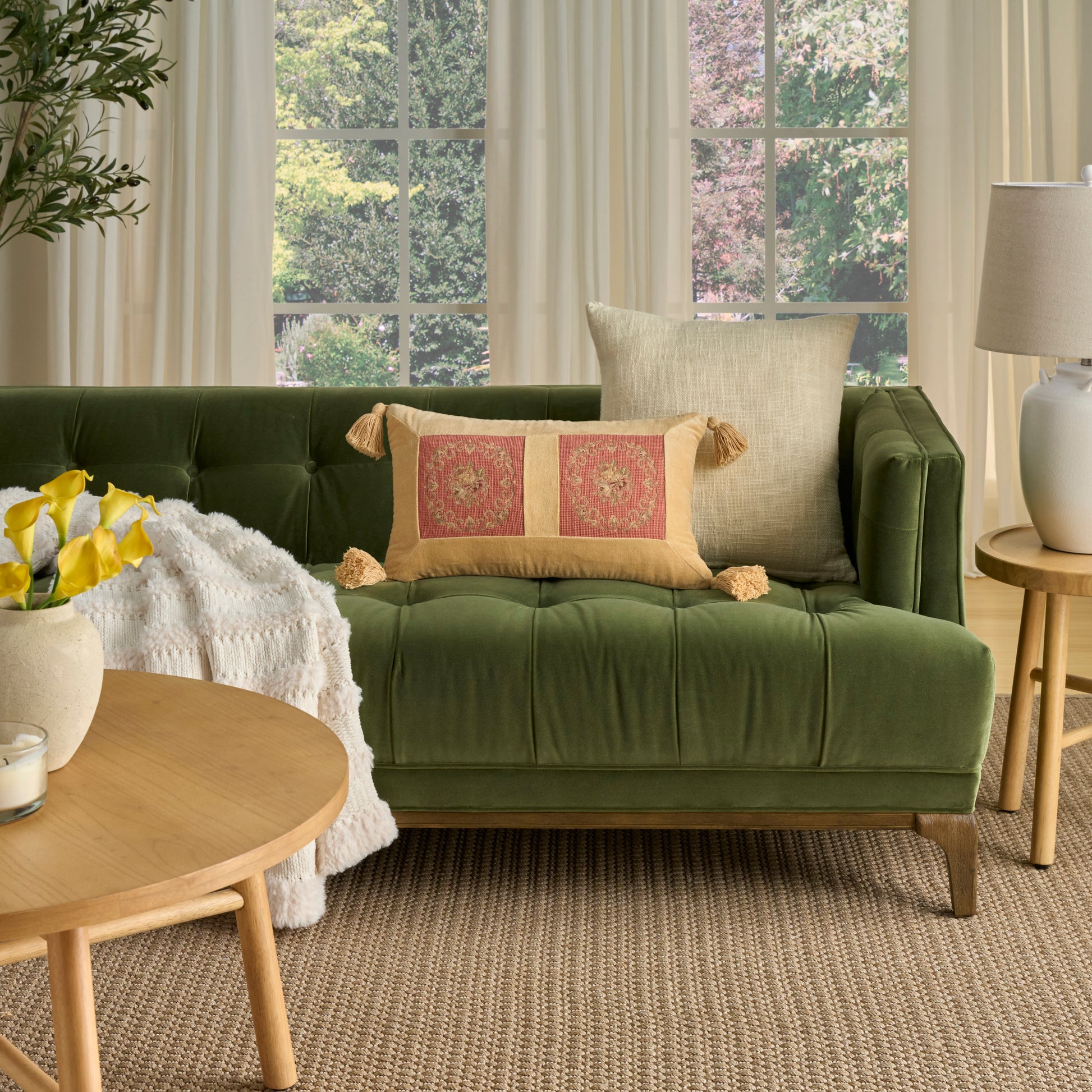 Olive green velvet tufted sofa with beige tassel pillows in a living room; round wood coffee table with yellow flowers.