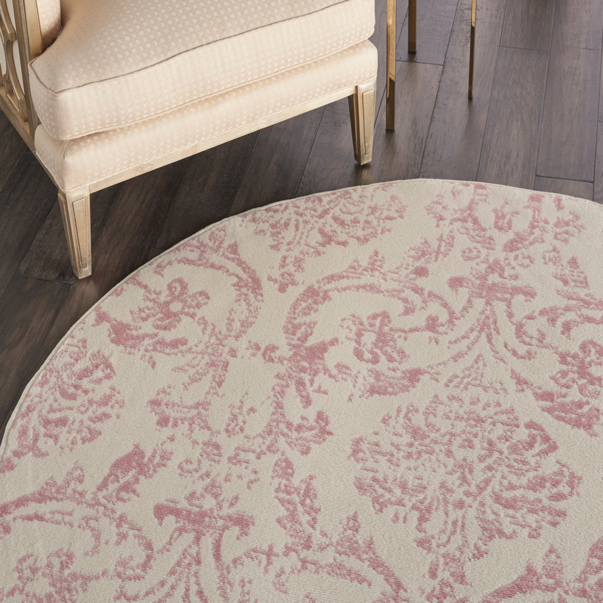 Round beige rug with pink damask floral pattern; paired with cream chair and dark wood floor.