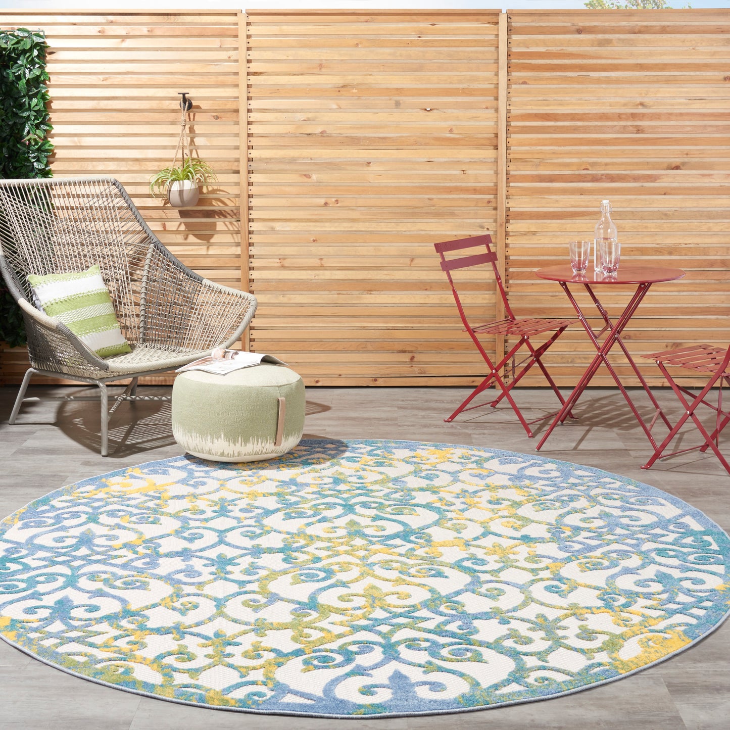 Outdoor patio with wooden fence, round blue-yellow patterned rug, wicker chair, green pouf, and red metal bistro table.