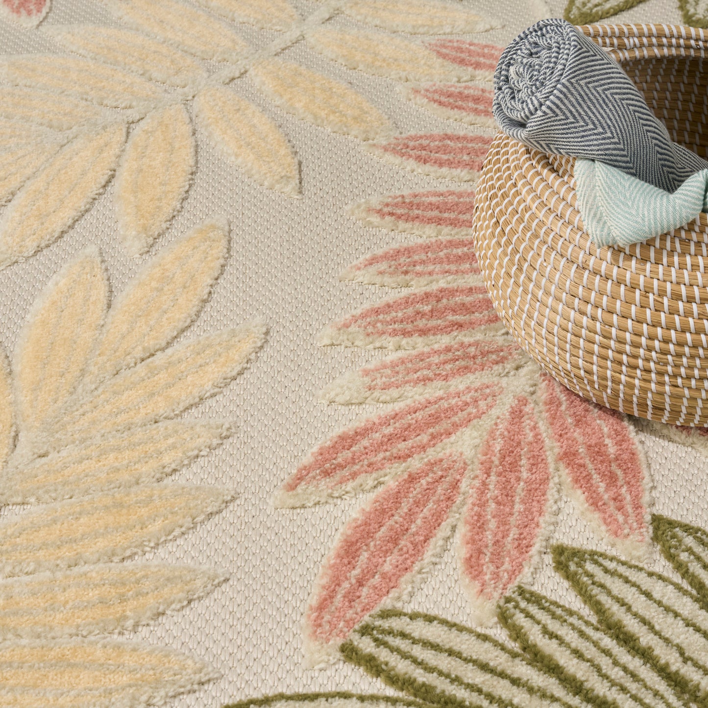 Round woven straw basket with rolled grey striped & pale blue cloth on a pastel floral rug with yellow & pink leaves.