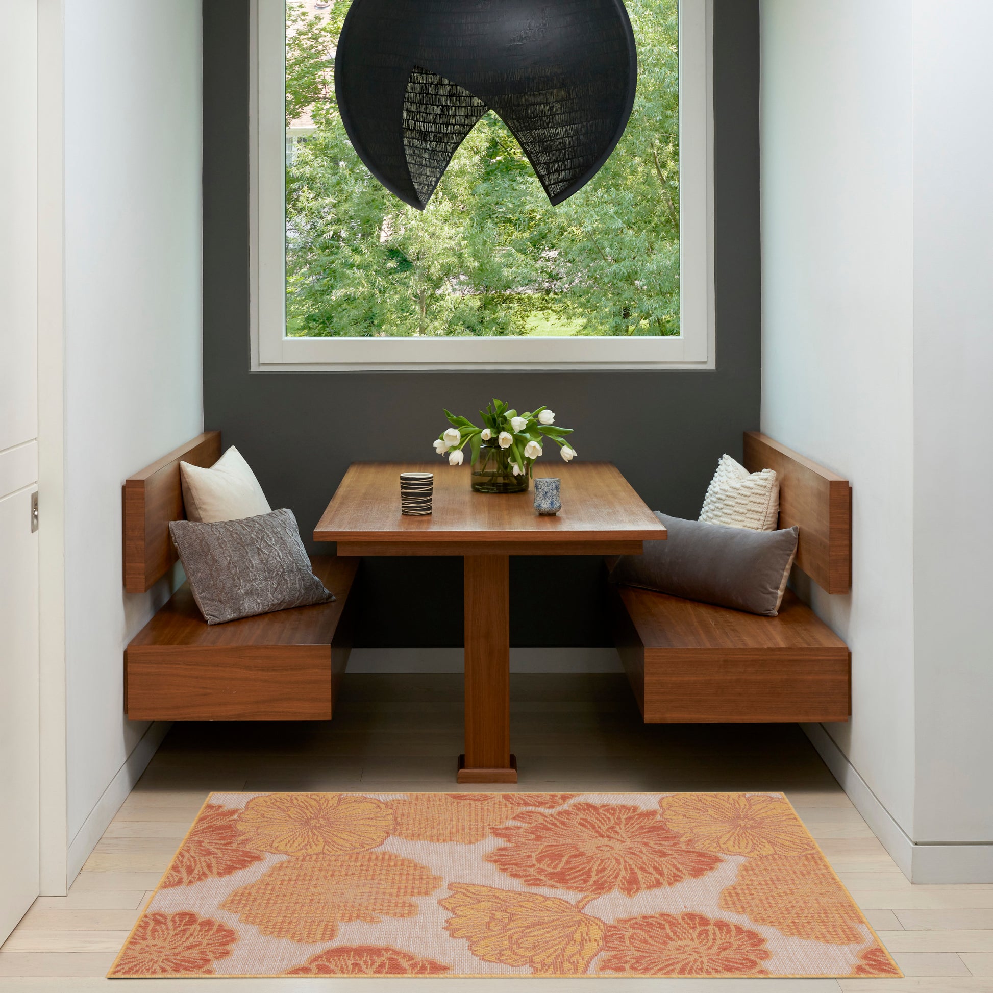 Wooden dining nook with L-shaped benches and square table, dark gray wall, white window, black pendant light, orange rug