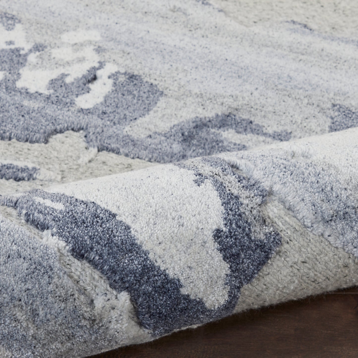 Soft plush rug in blue-gray and ivory with an abstract pattern and shaggy texture, folded on a dark wooden floor.