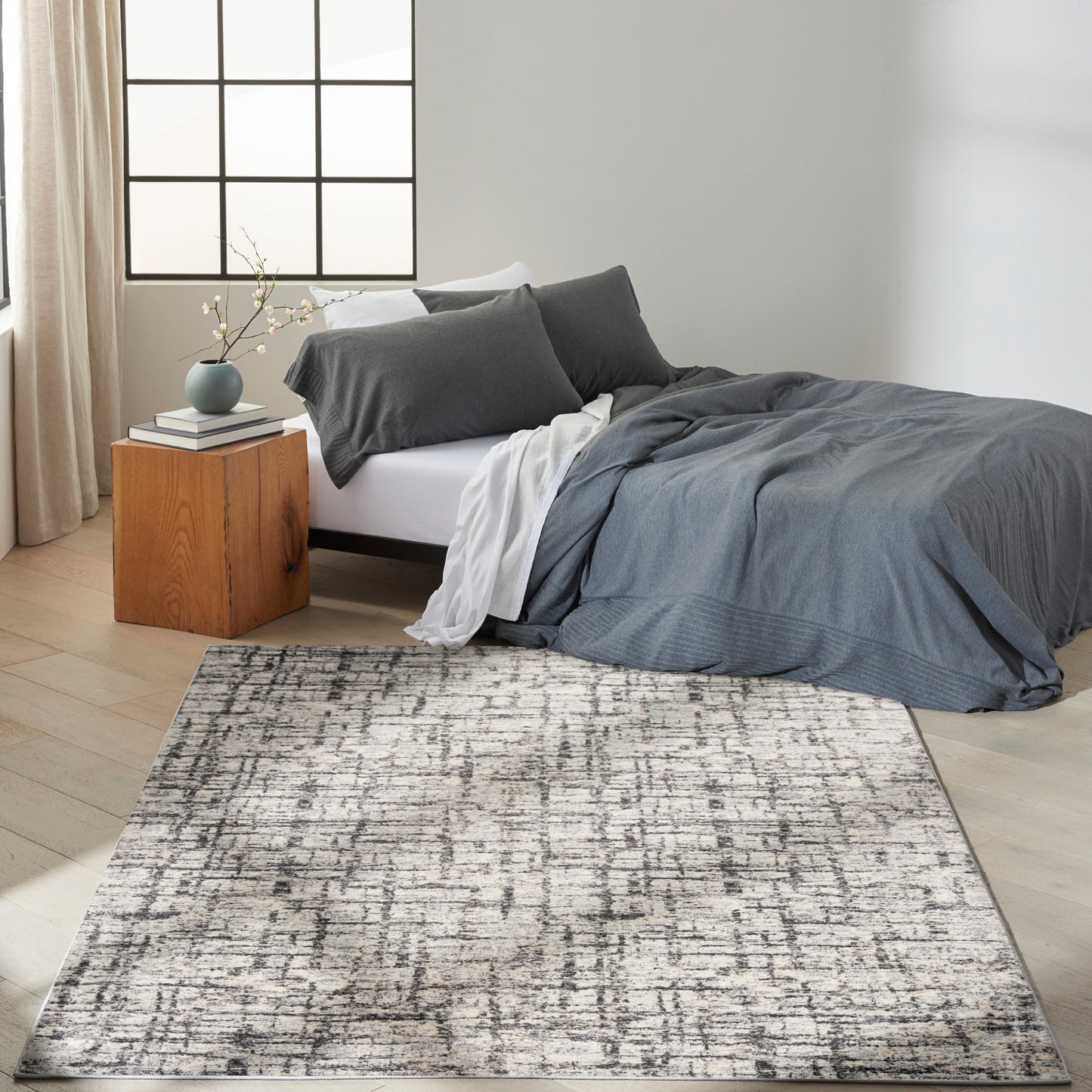 Minimalist bedroom with gray duvet and pillows, white sheets, wooden cube nightstand, vase of blossoms, and a textured rug.