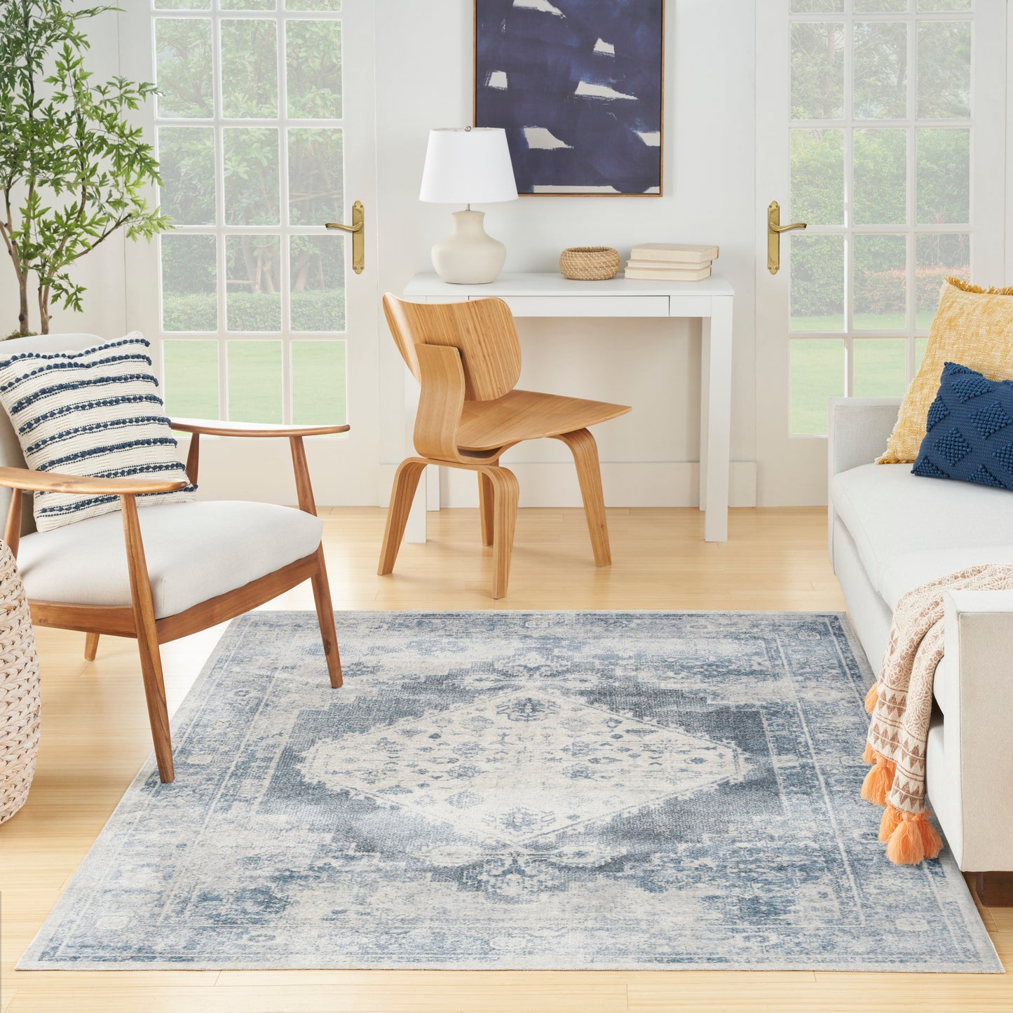 Light blue distressed area rug in a bright living room with mid-century chairs, white desk, plant, & blue-yellow cushions.