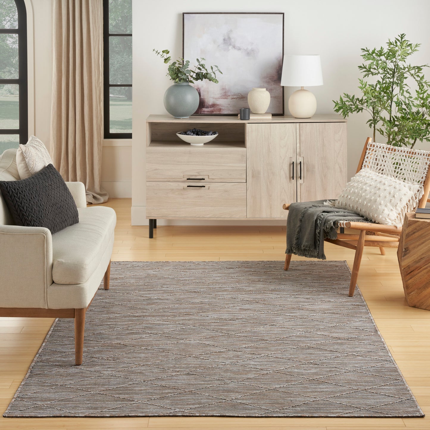 Light wood sideboard with drawers, abstract art, vases, lamp, plants, beige sofa, woven chair, and a gray patterned rug.