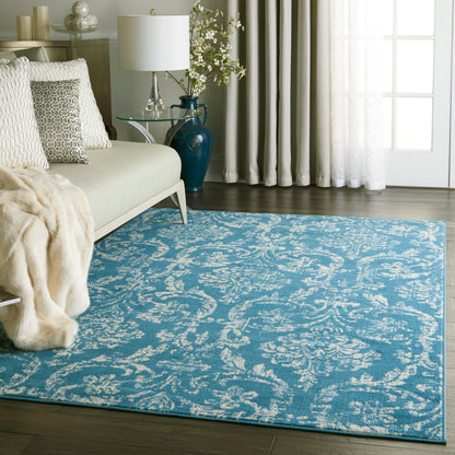 Cozy living room with teal-blue rug, cream sofa with textured pillows, glass lamp, blue vase, white curtains.