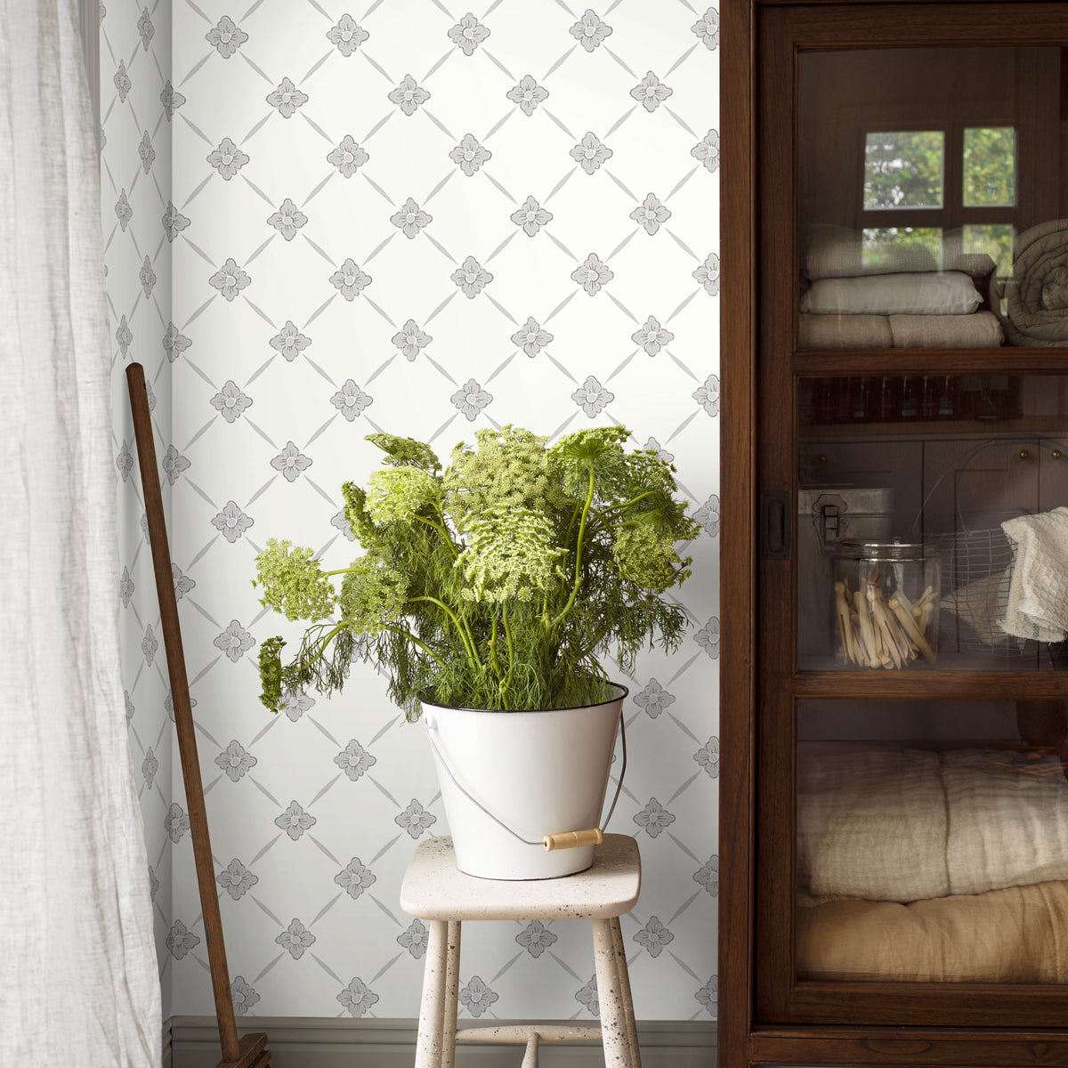 Green fern in a white metal bucket with a wooden handle on a small wooden stool; rustic decor with patterned wallpaper.