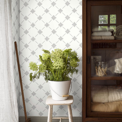Green fern in a white metal bucket with a wooden handle on a small wooden stool; rustic decor with patterned wallpaper.