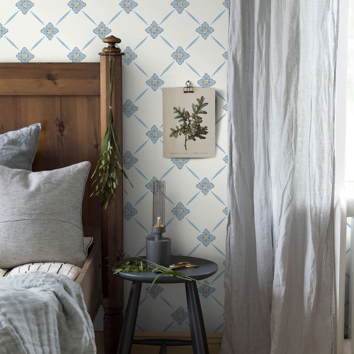 Cozy bedroom with wooden bed, blue floral lattice wallpaper, gray bedding, round table, dried herbs, botanical print, curtain.